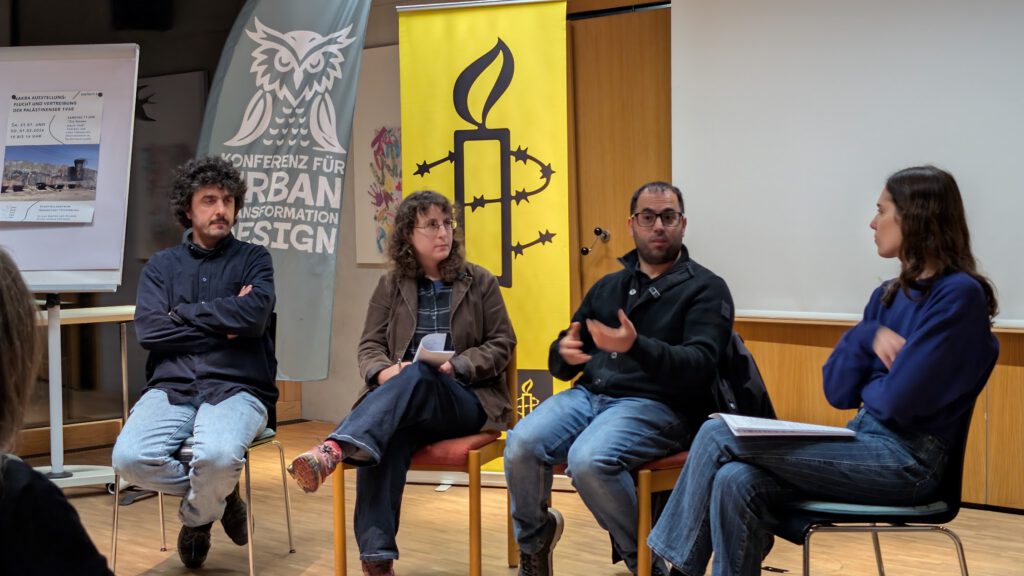 Vier Personen sitzen auf der Bühne und diskutieren, wobei ein Mann gestikuliert, während er spricht. Hinter ihnen sind Transparente angebracht, darunter eines mit einer Eule und ein weiteres mit einem gelben Amnesty International-Logo. An der Seite steht eine Staffelei mit Papieren.