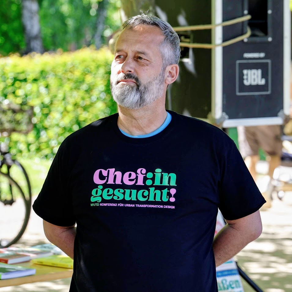 Uli Albicker. Ein Mann mit kurzen grauen Haaren und Bart steht im Freien und trägt ein schwarzes T-Shirt mit der Aufschrift "Chef:in gesucht!" in Pastellfarben. Im Hintergrund sind Grünpflanzen und ein Lautsprecher zu sehen.