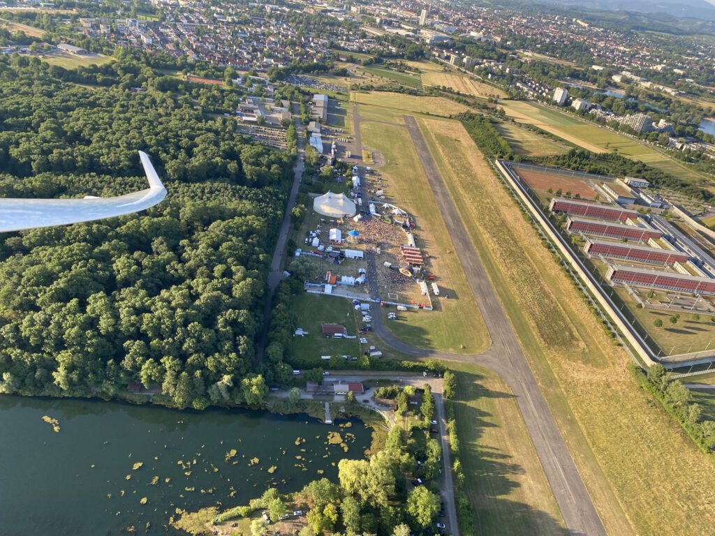 Luftaufnahme eines kleinen Flughafens mit Start- und Landebahn, Festzelten und Menschenmassen in der Nähe, grünen Bäumen, einem See und einer entfernten Stadt unter klarem Himmel, wobei ein Teil eines Flugzeugflügels zu sehen ist.