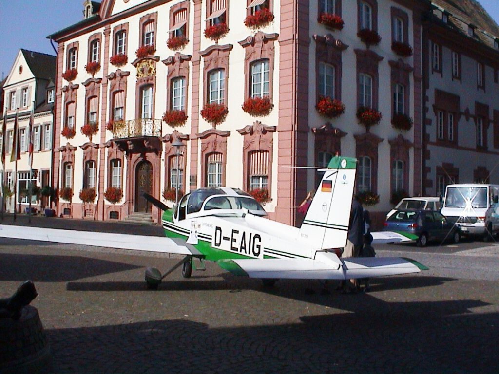 Ein kleines weiß-grünes Flugzeug parkt auf einem Platz mit Kopfsteinpflaster vor einem verzierten Gebäude mit roten Zierleisten und Blumenkästen an den Fenstern. Mehrere Autos sind in der Nähe geparkt.