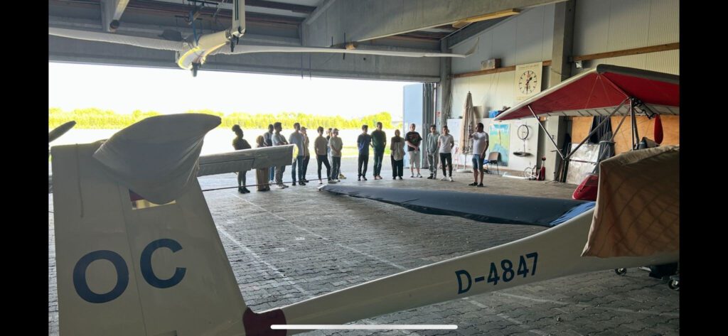 Eine Gruppe von Menschen steht in einer Reihe in einem offenen Hangar, mit einem Segelflugzeug mit der Kennzeichnung D-4847 und OC im Vordergrund. Sonnenlicht strömt von draußen herein und beleuchtet den Boden des Hangars.