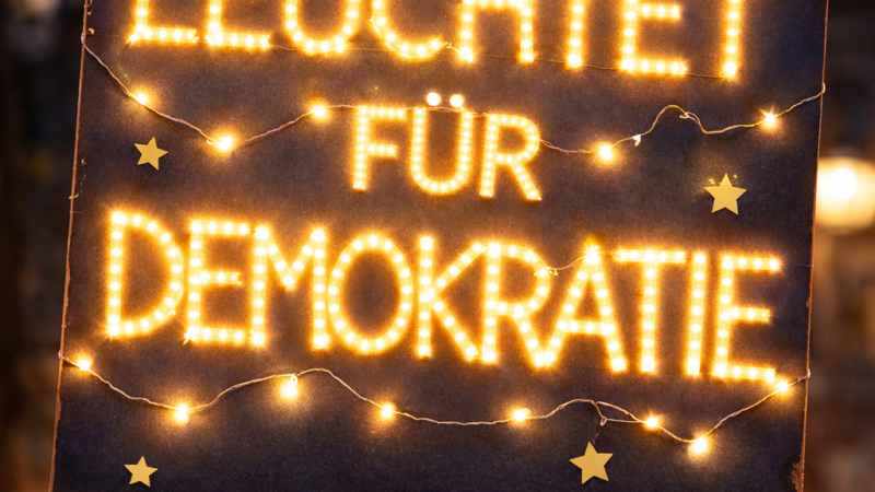 Eine Person hält während einer nächtlichen Versammlung ein mit gelben Lichtern beleuchtetes Schild mit der Aufschrift OFFENBURG LEUCHTET FÜR DEMOKRATIE, im Hintergrund sind unscharfe Menschen und Lichter zu sehen.