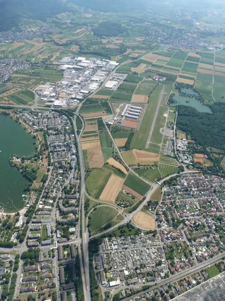 Luftaufnahme einer Stadt mit Wohnvierteln, Ackerland, einem großen See, einem Flughafen mit Start- und Landebahnen, Industriegebäuden und Straßen, die die verschiedenen Gebiete miteinander verbinden. Im Hintergrund sind grüne Hügel zu sehen.