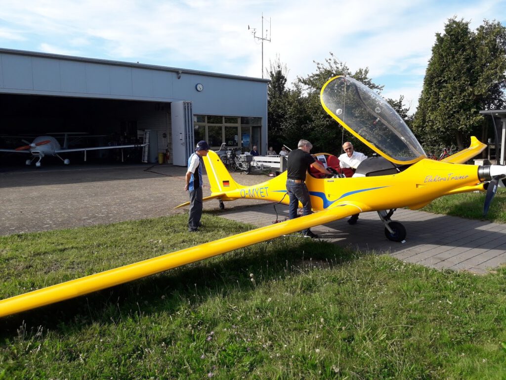 Drei Personen stehen in der Nähe eines leuchtend gelben Segelflugzeugs mit offener Cockpithaube vor einem Hangar. Ein weiteres Segelflugzeug ist im Inneren des Hangars zu sehen, und im Hintergrund stehen Bäume an einem sonnigen Tag.