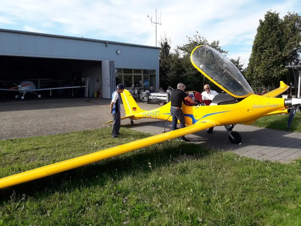 Drei Personen stehen in der Nähe eines leuchtend gelben Segelflugzeugs mit offener Cockpithaube vor einem Hangar. Ein weiteres Segelflugzeug ist im Inneren des Hangars zu sehen, und im Hintergrund stehen Bäume an einem sonnigen Tag.