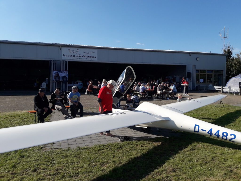 Ein weißes Segelflugzeug mit dem Kennzeichen D-4482 ist vor einem Hangar geparkt, in dem eine Gruppe von Menschen an einem sonnigen Tag im Freien sitzt und sich unterhält. Auf dem Hangarschild steht "Fliegergruppe Offenburg".