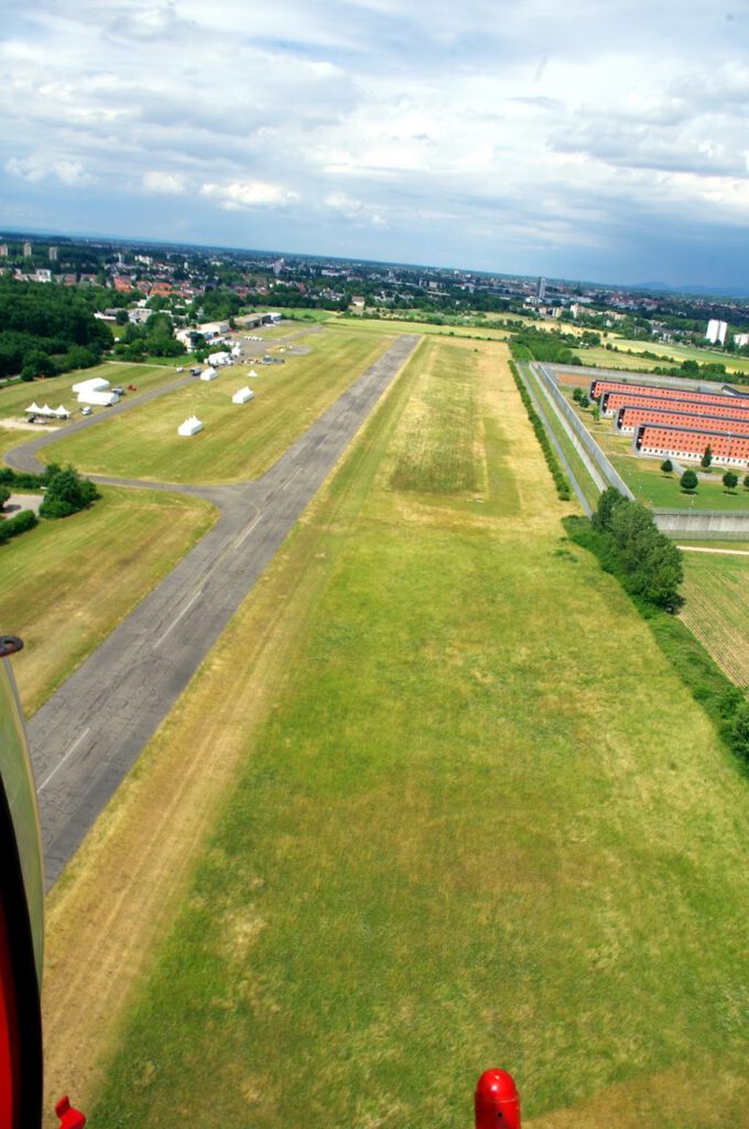 Luftaufnahme einer kleinen Start- und Landebahn eines Flughafens, umgeben von grasbewachsenen Feldern, einigen Hangars und nahegelegenen Gebäuden unter einem teilweise bewölkten Himmel.