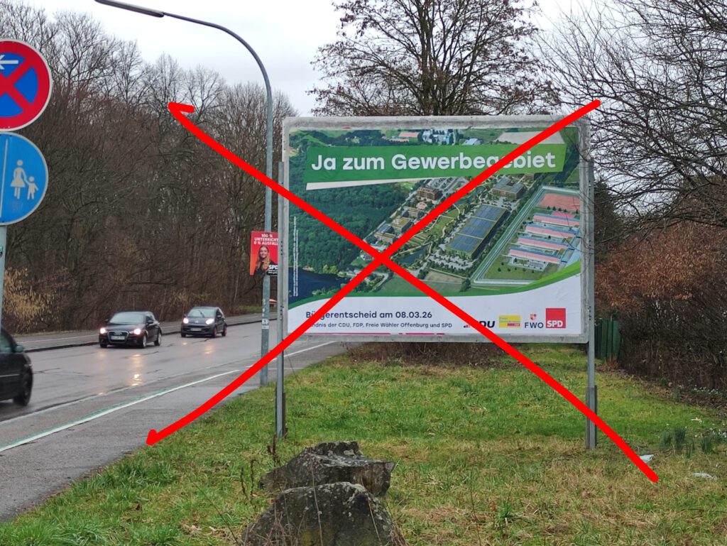 Eine Reklametafel am Straßenrand zeigt eine Luftaufnahme eines Industriegebiets mit dem deutschen Text "Ja zum Gewerbegebiet", der mit einem großen roten X durchgestrichen ist. Autos fahren auf der nassen Straße vorbei und Bäume säumen den Hintergrund.