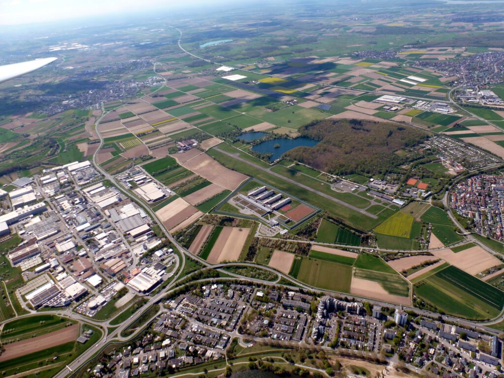 Luftaufnahme eines Flickenteppichs aus Ackerland, Fabriken, Wohngebäuden, Straßen und einem Waldgebiet mit einem Teich, alles umgeben von grünen Feldern und kleinen Städten unter einem klaren Himmel.