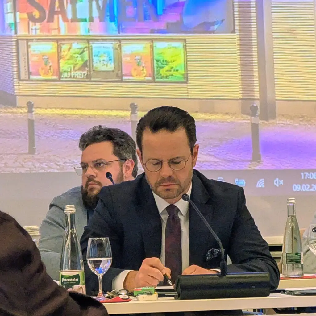 Zwei Männer in Anzügen sitzen an einem mit Wasserflaschen und Gläsern bedeckten Tisch, einer schreibt Notizen, der andere schaut nach vorne. Hinter ihnen ist eine bunte, beleuchtete Leinwand zu sehen.