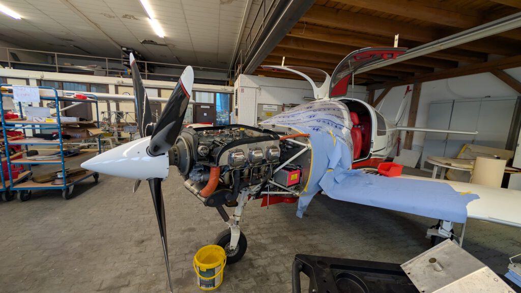 Ein kleines Flugzeug in einem Hangar, dessen Motorraum für Wartungsarbeiten geöffnet ist. Werkzeuge und Ausrüstung liegen in der Nähe, und eine blaue Folie bedeckt teilweise den Rumpf. Der Propeller und verschiedene Motorteile sind sichtbar.