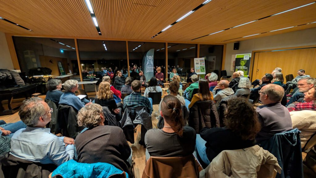 Eine Gruppe von Menschen sitzt im Kreis in einem hell erleuchteten Raum mit Holzwänden und -decke und hört einem Sprecher zu, der vorne neben Transparenten und großen Fenstern steht.