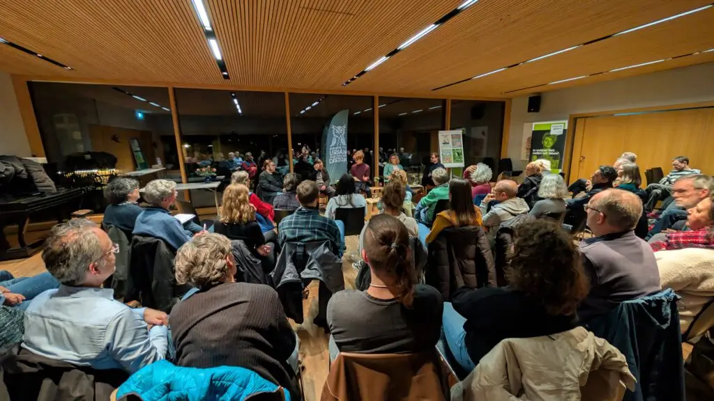 Eine Gruppe von Menschen sitzt im Kreis in einem hell erleuchteten Raum mit Holzwänden und -decke und hört einem Sprecher zu, der vorne neben Transparenten und großen Fenstern steht.