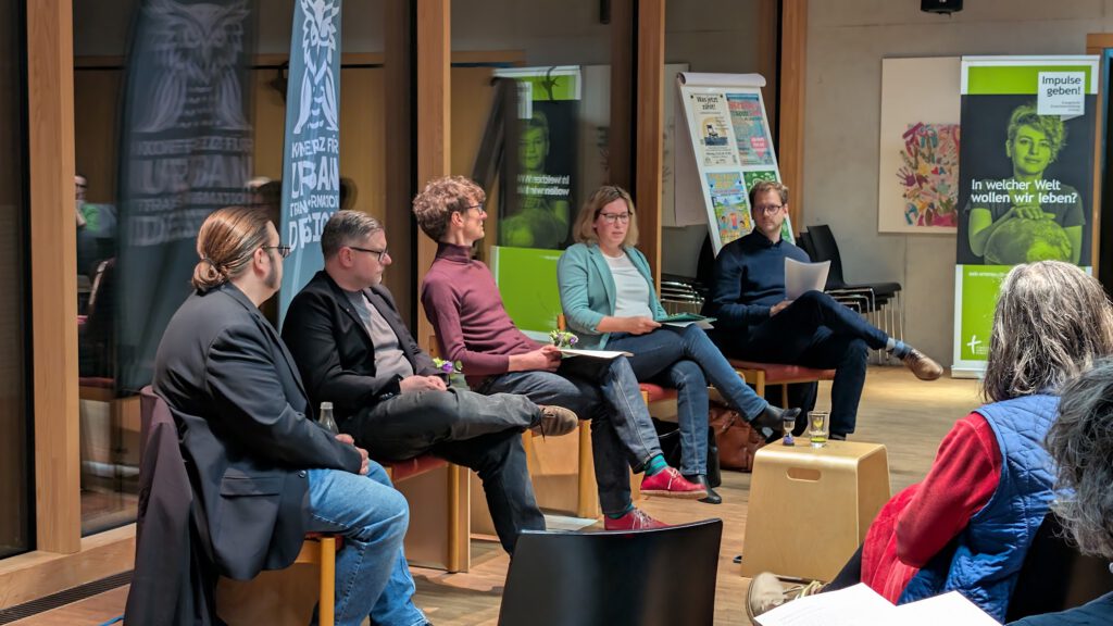 Fünf Personen sitzen in einem Halbkreis bei einer Podiumsdiskussion in einem modernen Raum, halten Papiere in der Hand und sprechen. Die Zuhörer sind im Vordergrund zu sehen. Im Hintergrund sind Plakate und Schaubilder zu sehen.