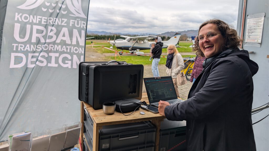 Eine Frau lächelt in die Kamera, während sie bei einer Veranstaltung im Freien an einem Laptop arbeitet. Hinter ihr sind ein kleines Flugzeug, Menschen und ein Banner mit der Aufschrift Konferenz für Urban Transformation Design zu sehen. Der Schauplatz scheint ein Flugplatz zu sein.