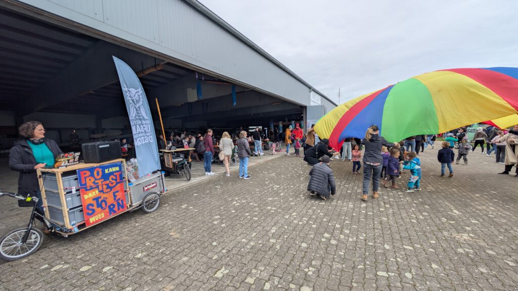 Menschen versammeln sich bei einer Gemeinschaftsveranstaltung im Freien in der Nähe eines großen offenen Gebäudes. Kinder spielen unter einem bunten Fallschirm, während Erwachsene zusehen oder sich unter die Leute mischen. Eine Frau steht an einem Wagen mit einem Protestschild und einem Transparent, und verschiedene Stände sind aufgebaut.