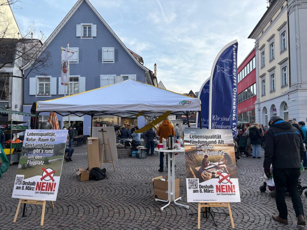 Eine Straßenszene zeigt einen Marktplatz mit einem weißen Zelt, um das sich Menschen versammelt haben, und Protestschildern in deutscher Sprache, die sich gegen ein Flughafenprojekt richten. Die Schilder sind mit roten Kreuzen und QR-Codes versehen, und nahe gelegene Gebäude säumen den Platz.