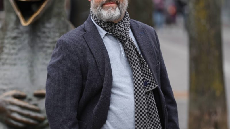 Ein Mann mittleren Alters mit grauem Bart steht im Freien und trägt einen Blazer, ein graues T-Shirt, einen Schal und eine braune Hose. Hinter ihm stehen eine große, skurrile Vogelstatue und mehrere Bäume an einer Stadtstraße.