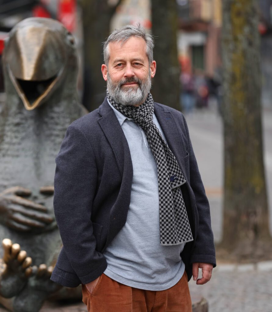 Ein Mann mittleren Alters mit grauem Bart steht im Freien und trägt einen Blazer, ein graues T-Shirt, einen Schal und eine braune Hose. Hinter ihm stehen eine große, skurrile Vogelstatue und mehrere Bäume an einer Stadtstraße.