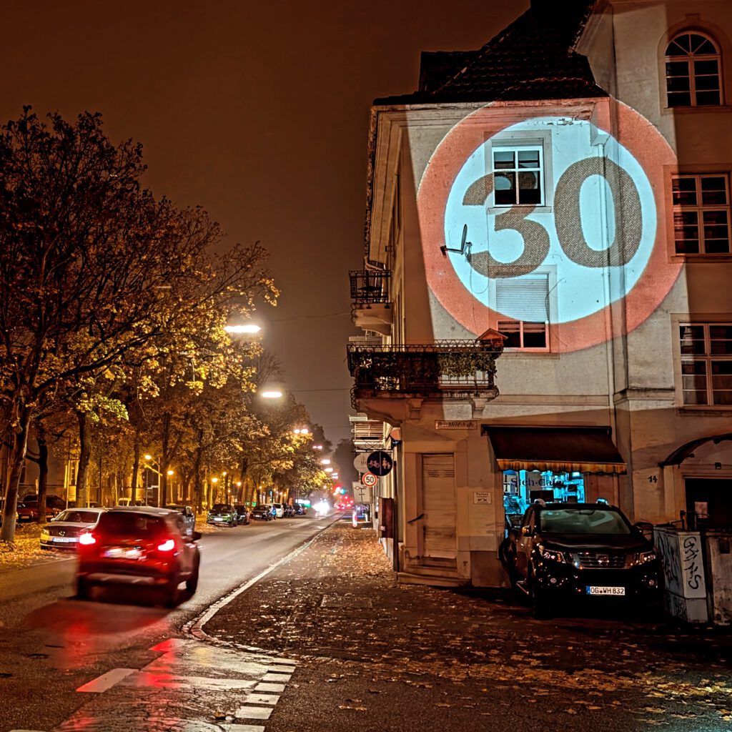 Eine nächtliche Stadtstraße mit vorbeifahrenden Autos; ein großes beleuchtetes Tempo 30-Schild ist auf die Seite eines Gebäudes projiziert. Der nasse Bürgersteig reflektiert die Straßenlaternen, und Herbstlaub säumt die Straße.