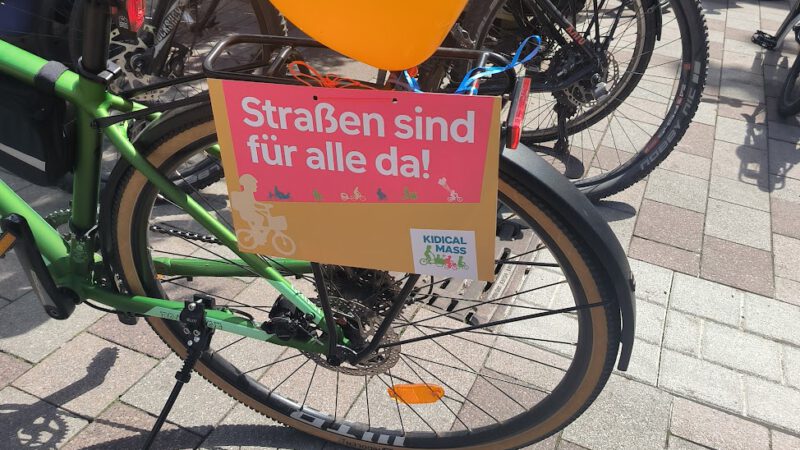 Ein grünes Fahrrad mit einem orangefarbenen Luftballon und einem Schild mit der Aufschrift Straßen sind für alle da! (Straßen sind für alle da!) ist bei einer Veranstaltung im Freien zwischen anderen Fahrrädern geparkt. Menschen in orangefarbenen Westen stehen in der Nähe einer Steinmauer.
