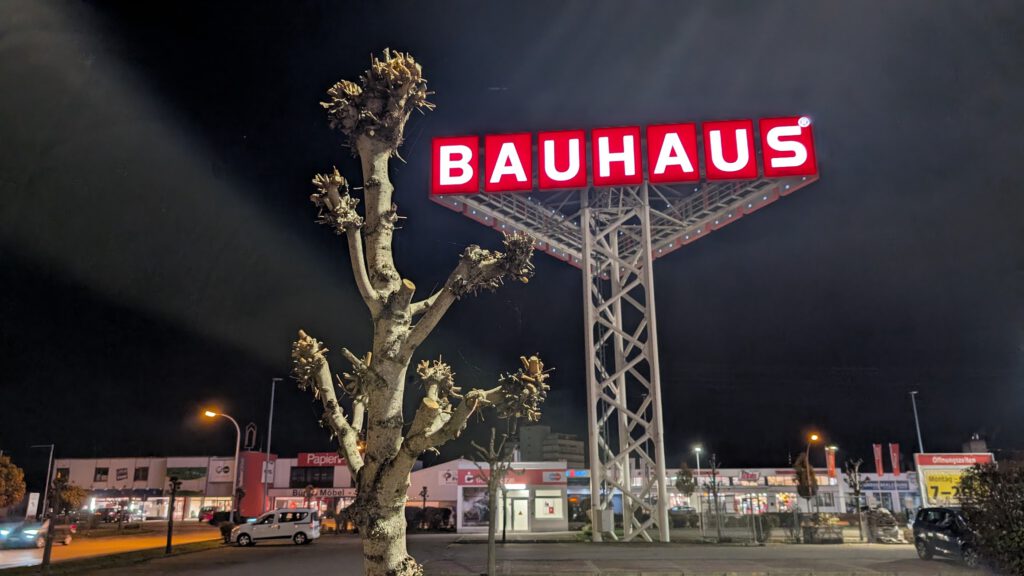 Ein blattloser, beschnittener Baum steht nachts vor einem großen beleuchteten BAUHAUS-Schild auf einer hohen Metallkonstruktion, mit einem gut beleuchteten Einkaufsbereich und geparkten Autos im Hintergrund.