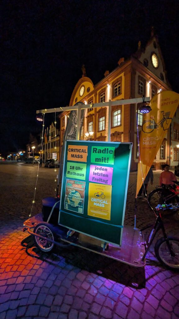 Ein Lastenfahrrad mit Critical Mass-Plakaten steht nachts auf einem Platz mit Kopfsteinpflaster vor einem hell erleuchteten historischen Gebäude mit Uhren und Giebeln. Banner und Lichter schmücken das Fahrrad und laden zur Teilnahme an der Veranstaltung ein.