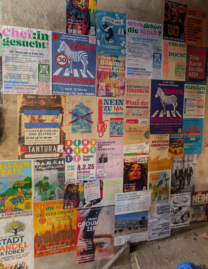 Eine Wand mit bunten Plakaten, die für Veranstaltungen, Proteste, Konzerte, politische Botschaften und Mitteilungen der Gemeinde werben, überlappen sich und sind in verschiedenen Schriftarten und Farben gehalten.