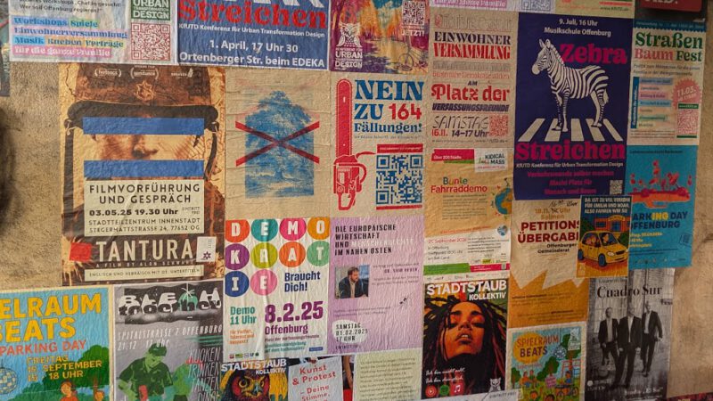 Eine Wand mit bunten Plakaten, die für Veranstaltungen, Proteste, Konzerte, politische Botschaften und Mitteilungen der Gemeinde werben, überlappen sich und sind in verschiedenen Schriftarten und Farben gehalten.