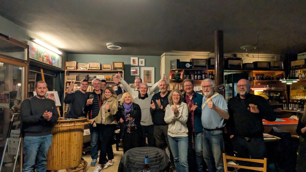 Eine Gruppe von Menschen steht in einer gemütlichen Bar oder einem Café zusammen, lächelt und hebt Getränke, was auf ein feierliches oder geselliges Beisammensein hindeutet. Regale mit Flaschen und verschiedene Dekorationen füllen den warm beleuchteten Raum.