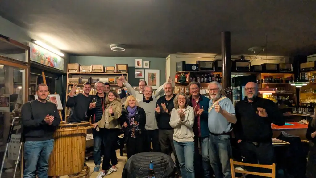 Eine Gruppe von Menschen steht in einer gemütlichen Bar oder einem Café zusammen, lächelt und hebt Getränke, was auf ein feierliches oder geselliges Beisammensein hindeutet. Regale mit Flaschen und verschiedene Dekorationen füllen den warm beleuchteten Raum.