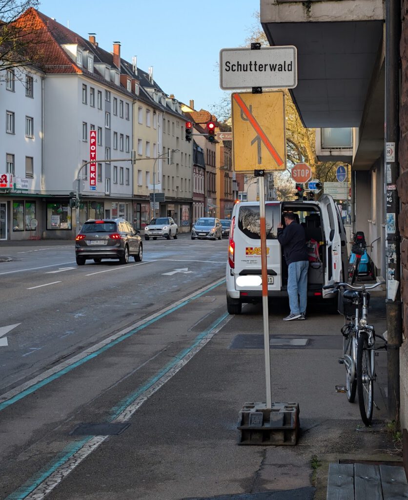Ein Mann steht am Heck eines weißen Lieferwagens, der auf einer Stadtstraße in der Nähe eines "Schutterwald"-Schildes geparkt ist. Ein Fahrrad ist auf dem Gehweg geparkt, und im Hintergrund fahren Autos an Geschäften und Wohnhäusern vorbei.