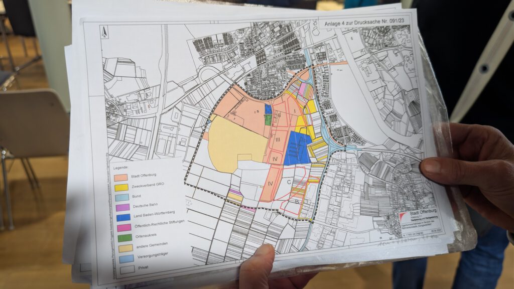 Eine Person hält einen gedruckten Stadtplan in der Hand, auf dem verschiedene Bereiche in unterschiedlichen Farben schattiert sind, um Zonen wie Wohngebiete, Gewerbegebiete und Industriegebiete zu kennzeichnen, wobei eine Legende und Straßennamen sichtbar sind.
