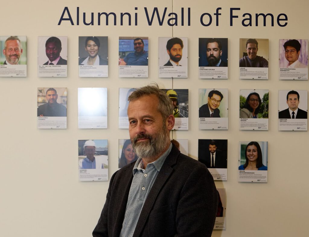 Ein Mann mit grauen Haaren und Bart steht vor einer Wand mit dem Titel Alumni Wall of Fame, auf der reihenweise Fotos und Kurzbiografien verschiedener Ehemaliger zu sehen sind.