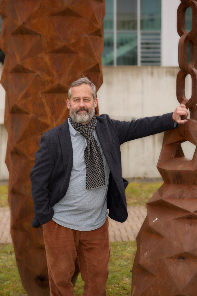 Ein bärtiger Mann, der ein graues Hemd, einen dunklen Blazer, einen gemusterten Schal und eine braune Hose trägt, lehnt im Freien an einer großen rostigen Metallskulptur und lächelt in die Kamera. Im Hintergrund ist ein modernes Gebäude zu sehen.