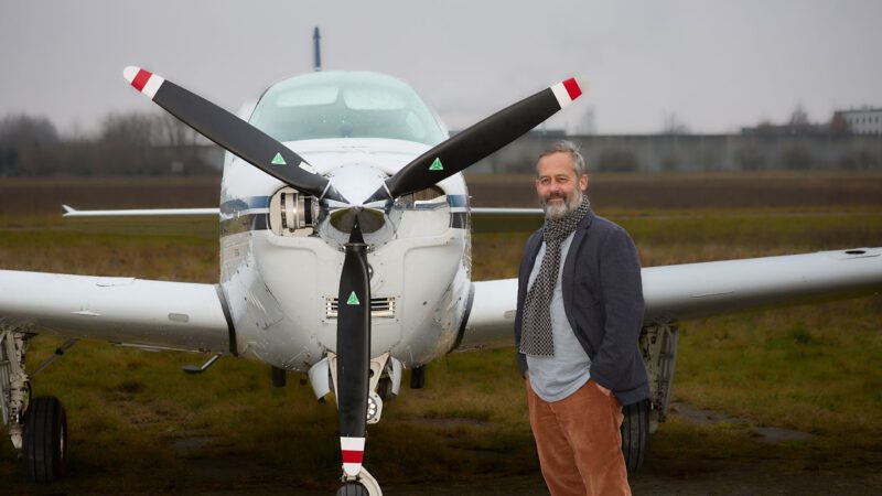 Ein Mann mit grauem Bart und legerer Kleidung steht lächelnd vor einem kleinen weißen Propellerflugzeug an einem bewölkten Tag, mit grasbewachsenen Feldern und Gebäuden im Hintergrund.