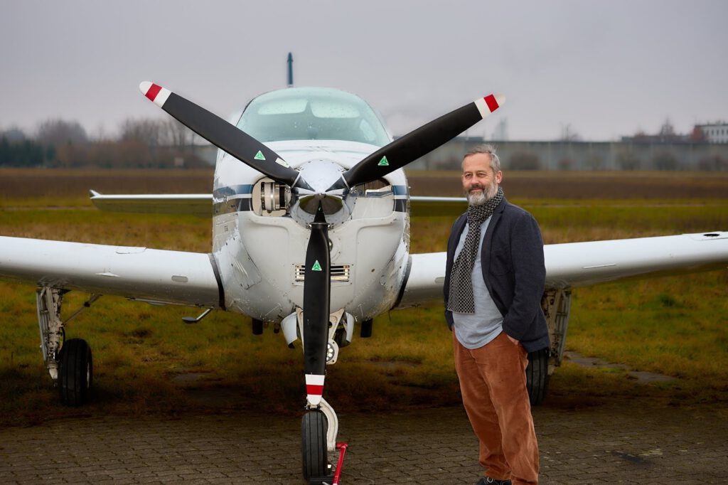 Ein Mann mit grauem Bart, Schal und brauner Hose steht an einem bewölkten Tag lächelnd neben einem kleinen weißen Propellerflugzeug, im Hintergrund sind Gras und Landebahn zu sehen.