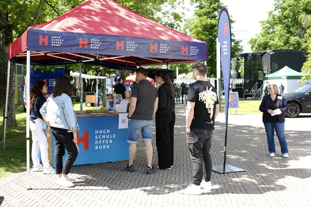 Bei einer Veranstaltung im Freien stehen Menschen an einem Informationsstand unter einem roten Zelt mit der Aufschrift Hochschule Offenburg. Bäume spenden Schatten, und auf dem Tisch sind verschiedene Displays und Broschüren zu sehen.