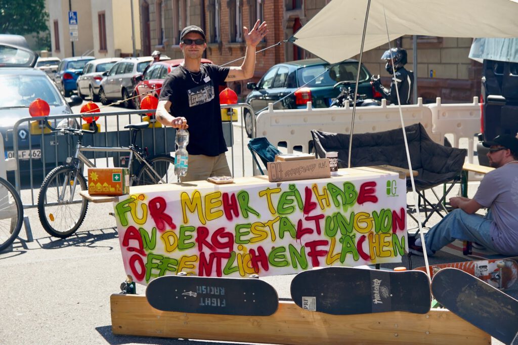Ein Mann steht hinter einem Tisch im Freien, winkt und lächelt. Auf dem Tisch stehen Skateboards und ein buntes Schild in deutscher Sprache, das für die Beteiligung der Öffentlichkeit an der Gestaltung von Freiräumen wirbt. Eine weitere Person sitzt in der Nähe, und im Hintergrund sind Fahrräder und Autos zu sehen.