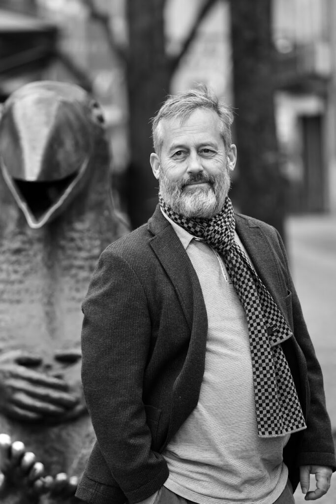 Ein bärtiger Mann mittleren Alters, der eine Jacke, ein T-Shirt und einen karierten Schal trägt, steht im Freien. Hinter ihm steht eine große Vogelskulptur mit offenem Schnabel, die leicht unscharf ist. Das Bild ist in schwarz-weiß gehalten.