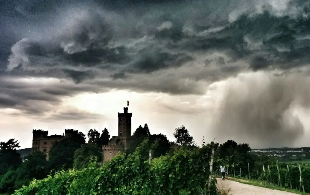Dunkle Gewitterwolken ziehen über Schloss Ortenberg auf, die von grünen Weinbergen umgeben ist, während im Vordergrund eine Person einen Weg entlangläuft. Die Atmosphäre ist dramatisch und ahnungsvoll.