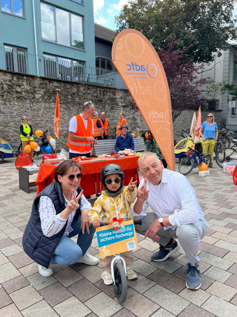 Ein kleines Kind mit Sonnenbrille und Helm posiert mit zwei Erwachsenen, die alle Friedenszeichen machen, bei einer Veranstaltung im Freien mit Fahrrädern, Luftballons und Menschen in Sicherheitswesten. Das Kind hält ein Schild auf Deutsch über sichere Radwege.