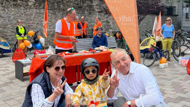 Ein kleines Kind mit Sonnenbrille und Helm posiert mit zwei Erwachsenen, die alle Friedenszeichen machen, bei einer Veranstaltung im Freien mit Fahrrädern, Luftballons und Menschen in Sicherheitswesten. Das Kind hält ein Schild auf Deutsch über sichere Radwege.