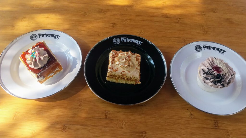 Drei verschiedene Torten, serviert auf weißen und schwarzen Tellern, die in einer Reihe auf einem Holztisch angeordnet sind, jede mit einzigartigen Belägen und Dekorationen, wobei das Sonnenlicht sanfte Schatten wirft.