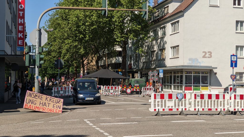 Eine Stadtstraße ist teilweise durch weiße und rote Absperrungen blockiert. Auf einem Schild in deutscher Sprache steht "UNSERE KREUZUNG HAT EIN BAUMPROBLEM". Menschen stehen in der Nähe von Bäumen und einem schwarzen Lieferwagen unter einem Zelt. Gebäude und geparkte Autos sind zu sehen.