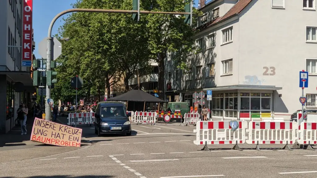 Eine Stadtstraße ist teilweise durch weiße und rote Absperrungen blockiert. Auf einem Schild in deutscher Sprache steht "UNSERE KREUZUNG HAT EIN BAUMPROBLEM". Menschen stehen in der Nähe von Bäumen und einem schwarzen Lieferwagen unter einem Zelt. Gebäude und geparkte Autos sind zu sehen.