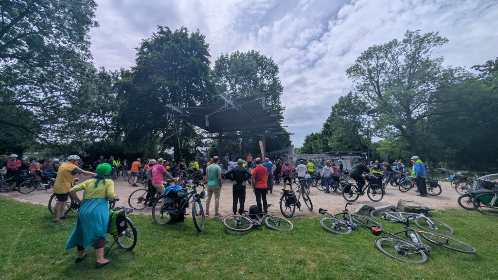 Group of cyclists with helmets gathered in a park near a shaded stage, many bikes parked on the grass.