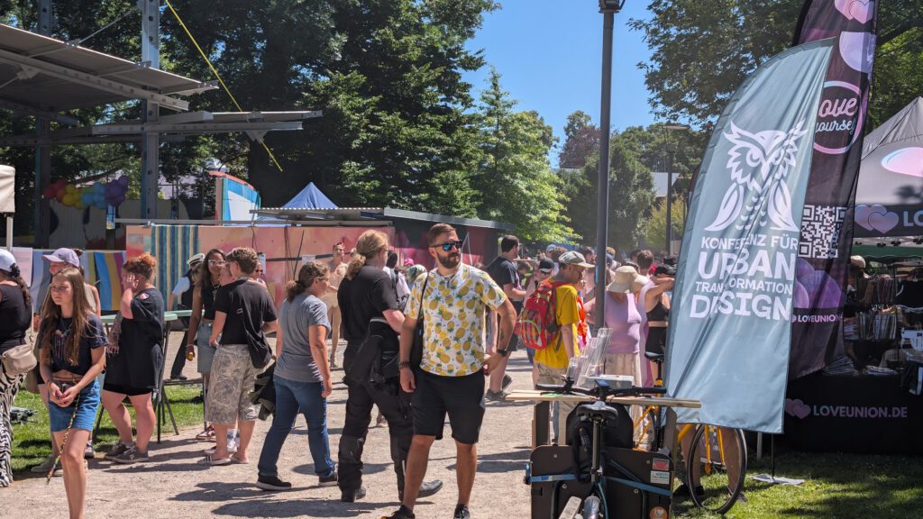 Menschen schlendern durch ein lebhaftes Freiluftfestival mit Ständen, Bannern und Kunstausstellungen. Auf einem blauen Transparent steht "Konferenz für Urban Transformation Design". Bäume und Sonnenschein schaffen eine lebendige Sommeratmosphäre.