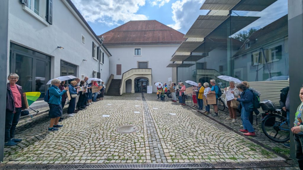 Eine Gruppe von Menschen steht sich in zwei Reihen in einem gepflasterten Innenhof gegenüber, einige halten Schilder und Regenschirme. Die Kulisse liegt zwischen modernen Glas- und traditionellen weißen Gebäuden unter einem teilweise bewölkten Himmel.