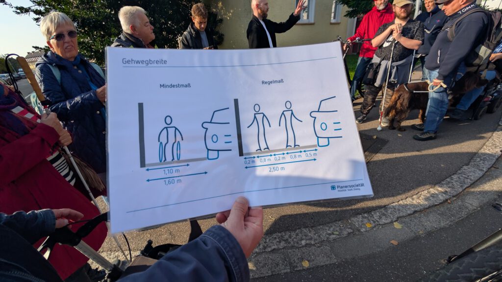 Eine Person hält ein Plakat in der Hand, auf dem die Richtlinien für die Breite deutscher Bürgersteige mit Diagrammen von Personen und Autos dargestellt sind. Mehrere Personen und ein Hund sind draußen versammelt, einige mit Mobilitätshilfen.